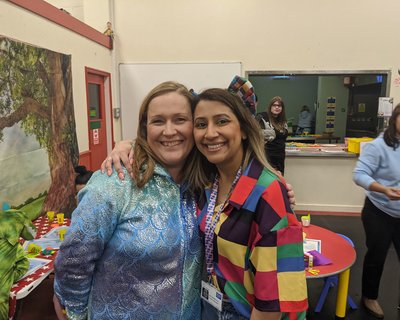 Two women hug each other whilst smiling and looking at the camera. One is wearing a metallic turquoise coloured top and the other is wearing a multi-coloured patch top. They are in a building with other people behind them.