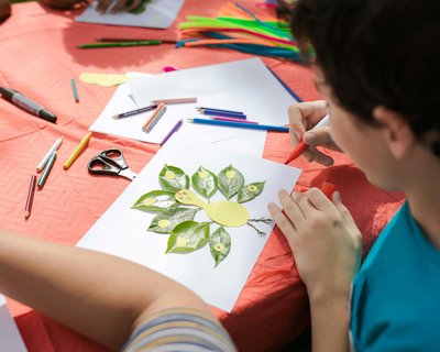 A child is sitting at a table covered with an orange tablecloth, engaging in an art project. The child is working on a piece of paper that has a collage of leaves and other materials arranged to form the shape of a bird.