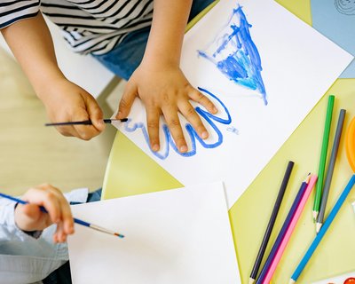 Two children are painting at a table. One child is outlining their hand in blue on a white sheet of paper, while the other is using a paintbrush on a separate piece. The table is covered with art supplies, including coloure pencils, markers, paintbrushes.