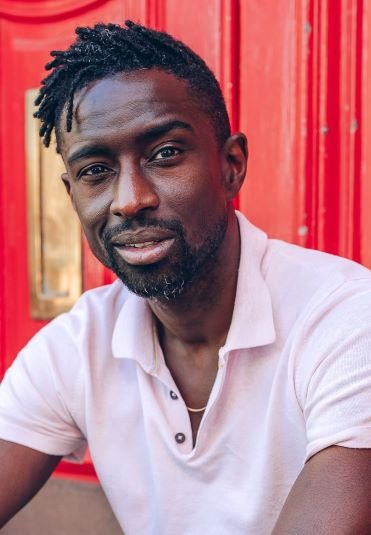 A headshot of Jeffrey Boakye. He looking straight at the camera, leaning forward with a small smile, in front of a bright red background.