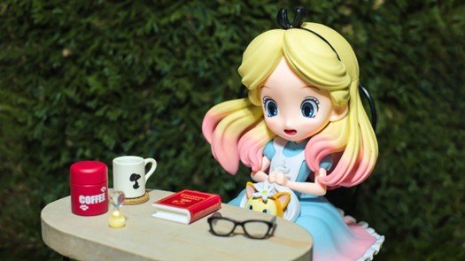An Alice in Wonderland doll in a blue dress with a model cup of coffee and a book