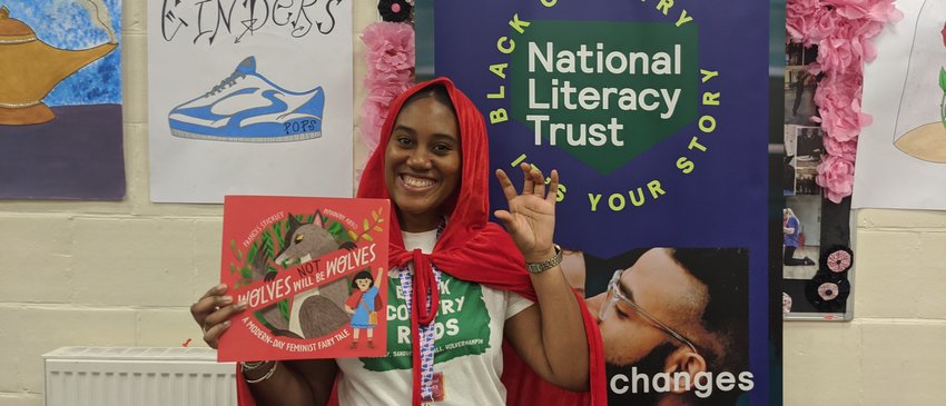 A person dressed in a red cape is holding a book titled "Wolves Will Be Wolves" in front of a banner for the Black Country National Literacy Trust. The background includes artwork of a shoe labelled "Cinders" and other decorations.