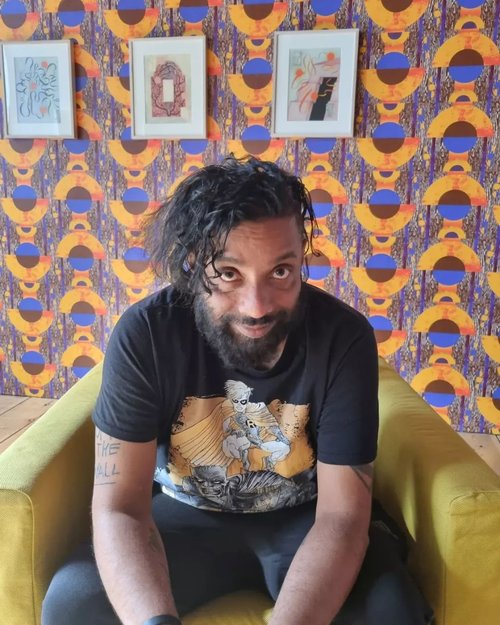 A head shot of Bobby Joseph. He is smiling at the camera, sitting forward resting his elbows on his knees. He is on a bright yellow chair and there is brightly coloured patterned wallpaper behind him.
