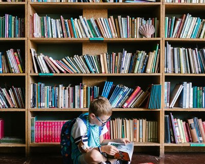 A young boy with glasses and a backpack sits on the floor in front of a large wooden bookshelf filled with books. He is reading a book, with several other books scattered around him. The bookshelf has multiple shelves containing books of various sizes and