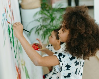 Two children are painting on a large white canvas. The child in the foreground has curly hair and is holding a paintbrush in one hand and a small container of red paint in the other. The child in the background is partially obscured but also appears to be